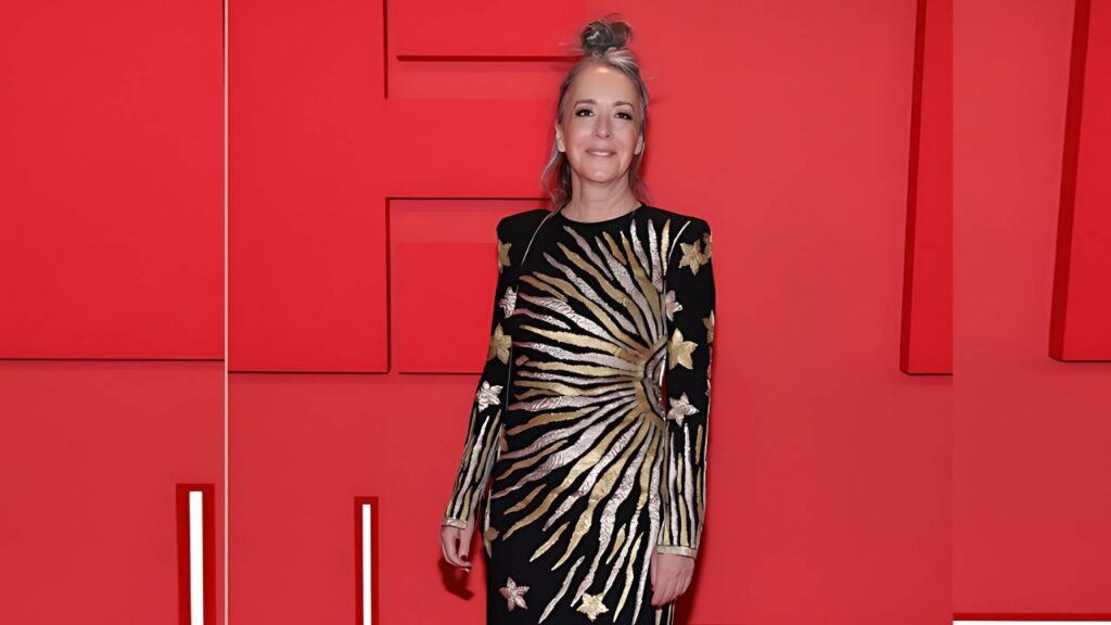 Jane Adams in a black long-sleeve gown with gold sunburst and star details poses against a bold red geometric backdrop.