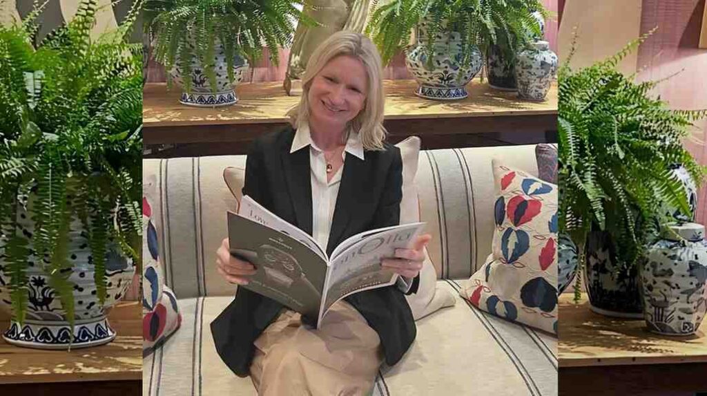 Jane Alexander in a black blazer sits on a striped sofa, reading a magazine, with ferns in blue pots behind her.