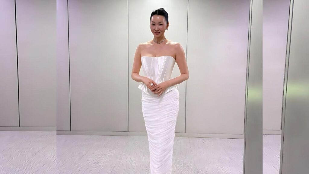 Jang Yoon-ju in a strapless white gown with a ruched skirt stands in a sleek, metal-panel room, hands clasped and smiling.