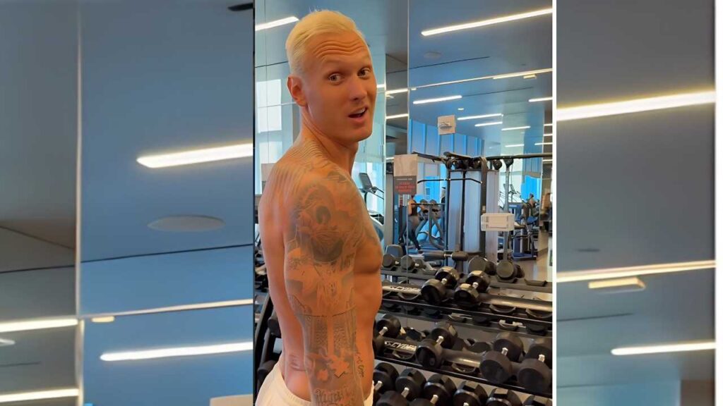 Janis Timma with blonde hair turns over his shoulder in a gym, revealing back tattoos amid dumbbells and blue walls.