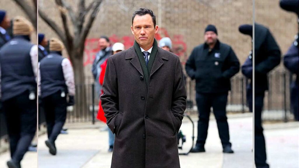 Jeffrey Donovan in a brown overcoat and green scarf stands with hands in pockets on a city sidewalk, blurred security guards behind.