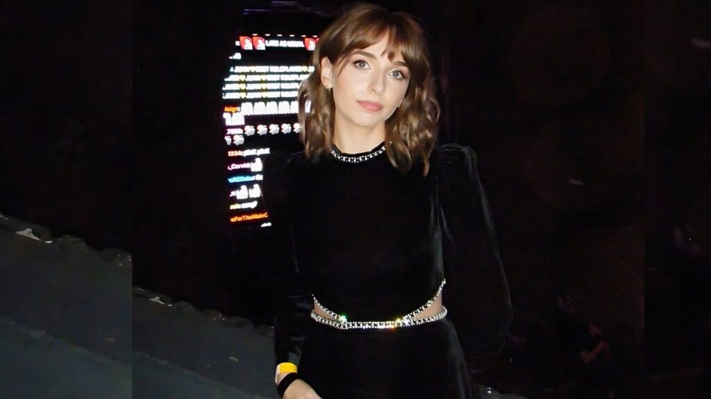 Jenn McAllister in a black velvet dress with a jeweled belt and studded choker poses before a dark backdrop, neon screen behind.