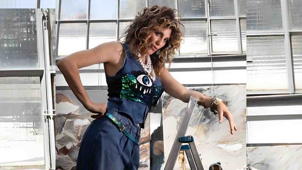 Jennifer Esposito in a blue denim jumpsuit leans on a step ladder, hand on hip, pearls, sequined eye graphic on her top in an airy studio.