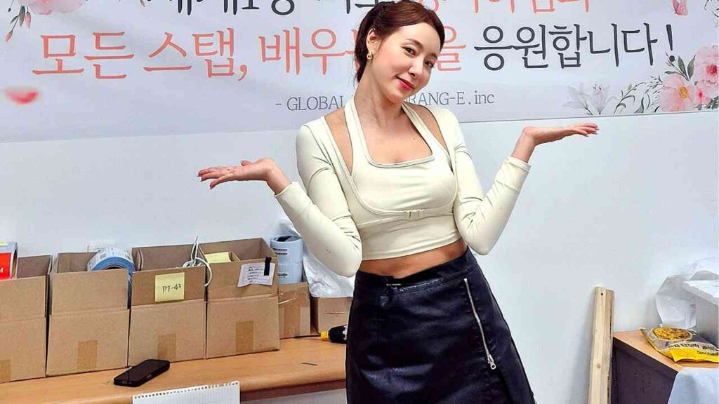 Jeong Seo-ha (정서하) in cream cropped top and black leather skirt poses with arms out, in front of boxes and a floral banner.