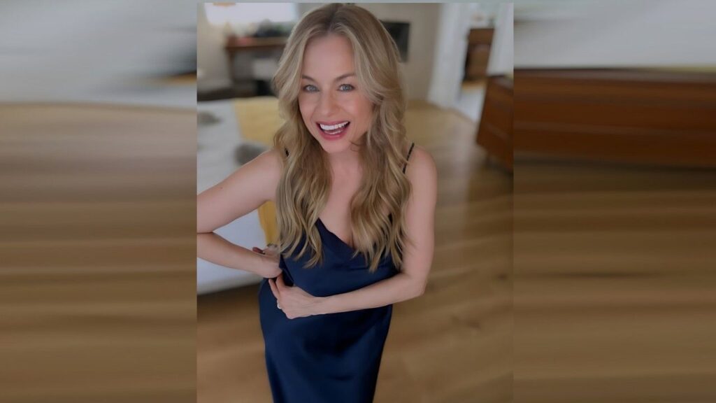 Jessica Collins in a navy dress poses indoors on a wooden floor, hands on her waist, blurred living room behind.