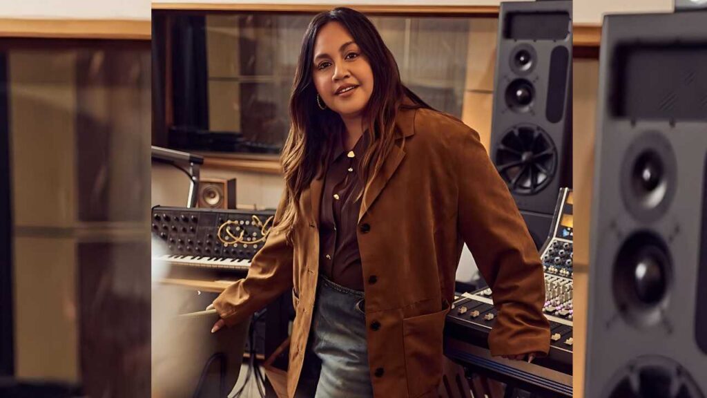 Jessica Mauboy with long hair in a brown suede coat leans on a music studio console amid warm lighting and large speakers.