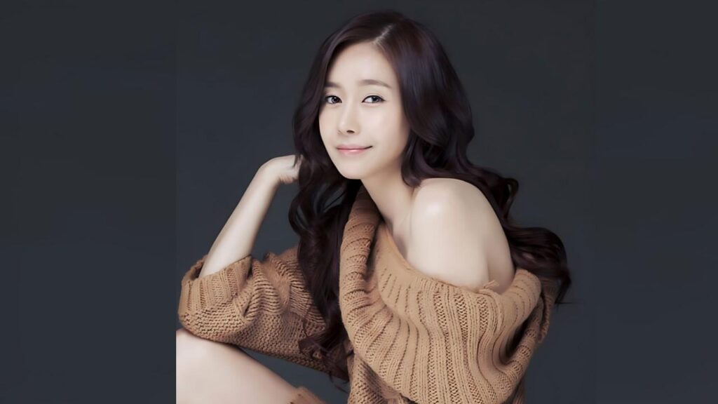 Ji Seo-yoon with long, wavy hair wears a tan knit off-shoulder sweater, resting her chin on her hand against a gray backdrop.