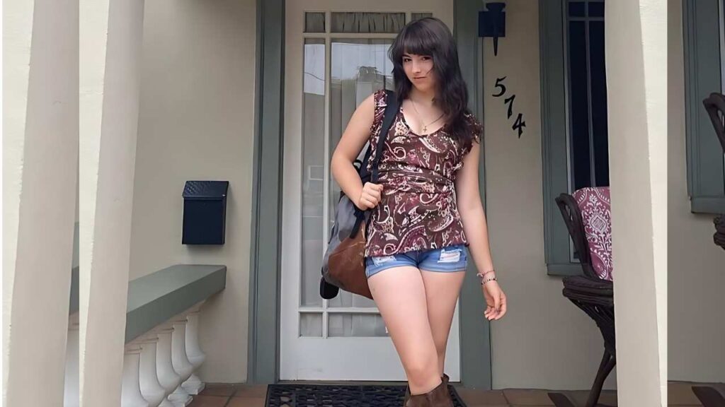 Jillian Armenante with shoulder-length dark hair stands on a porch in a paisley top and denim shorts, carrying a brown shoulder bag.