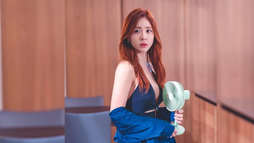 Jin Ye-sol with long auburn hair in a blue halter top and jacket draped off her shoulders, holding a mint handheld fan.
