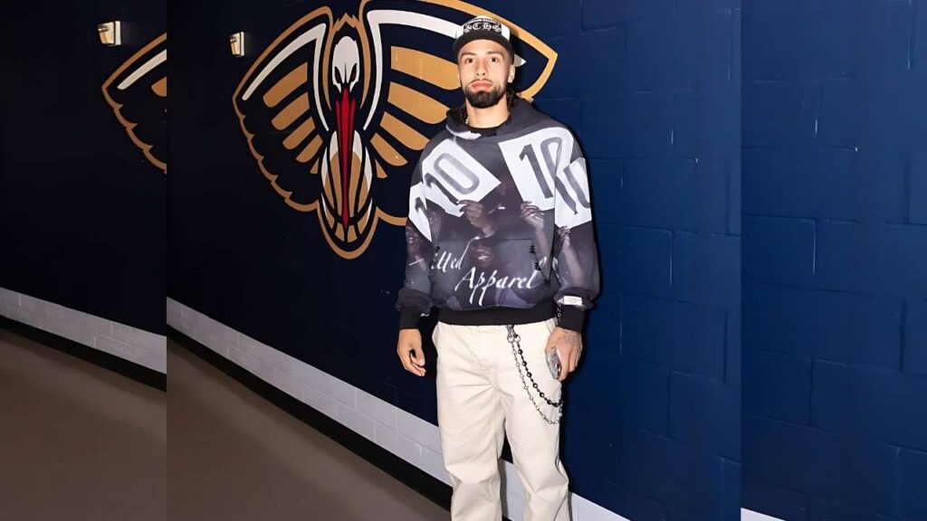 Jose Alvarado in an oversized graphic hoodie and beige pants with a chain, standing in a blue hallway beside a large pelican mural, holding a phone.