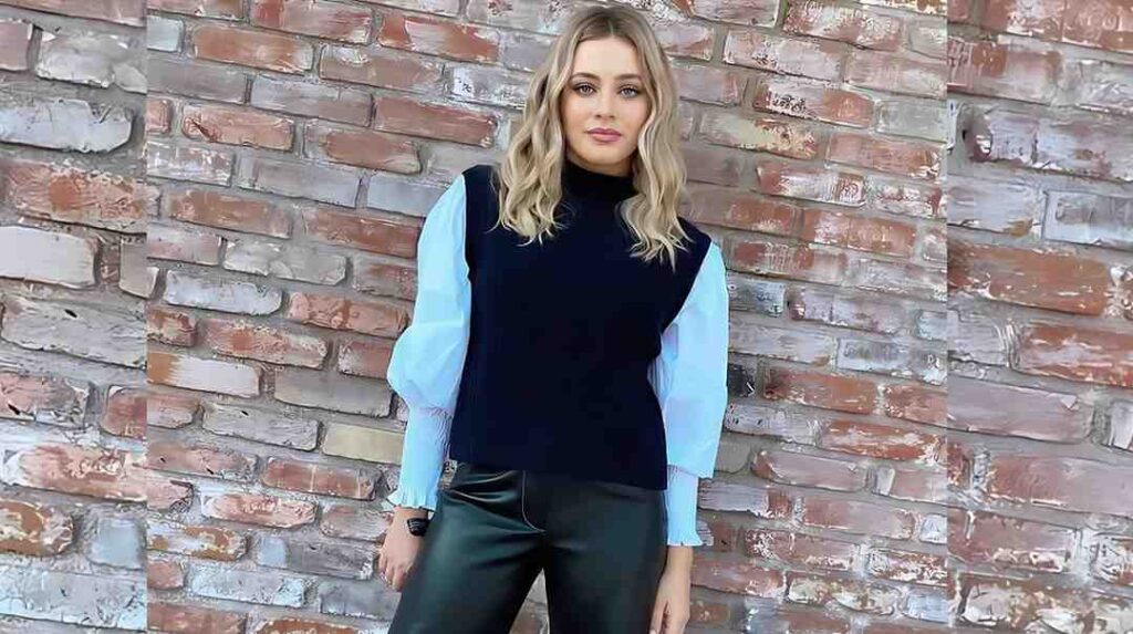 Josephine Langford with wavy blonde hair poses before a brick wall, wearing a light-blue puff-sleeve blouse under a black sleeveless top and black pants.
