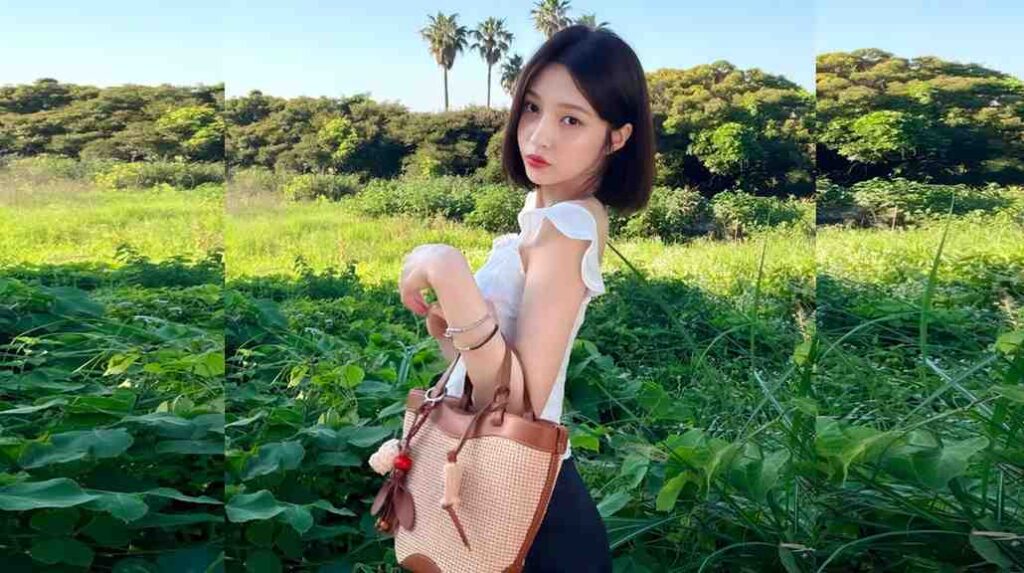 Joy in a white top with ruffled sleeves stands in a sunlit green field, holding a woven straw bag.