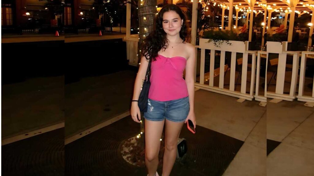 Julia Antonelli in a pink top and denim shorts poses at night, smiling, with a black purse as lights glow behind white railing.