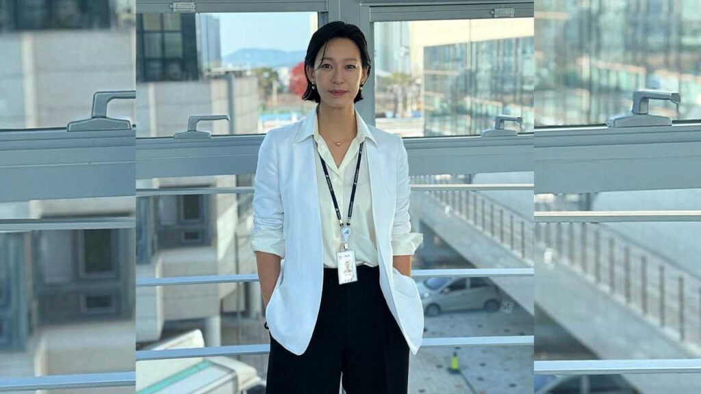 Jung Ae-yun (정애연) in white blazer and cream blouse stands on a glass balcony, hands in pockets, ID badge on a lanyard, urban skyline behind.