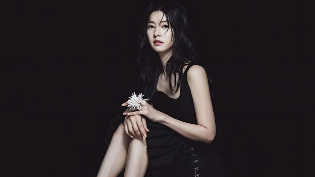 Jung So-min in black dress sits against a dark backdrop, holding a white spiky flower; long black hair frames her face.
