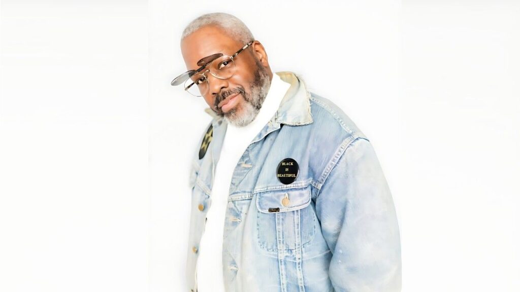 Kadeem Hardison in a light denim jacket and white shirt, round glasses, wearing a 'BLACK IS BEAUTIFUL' badge.