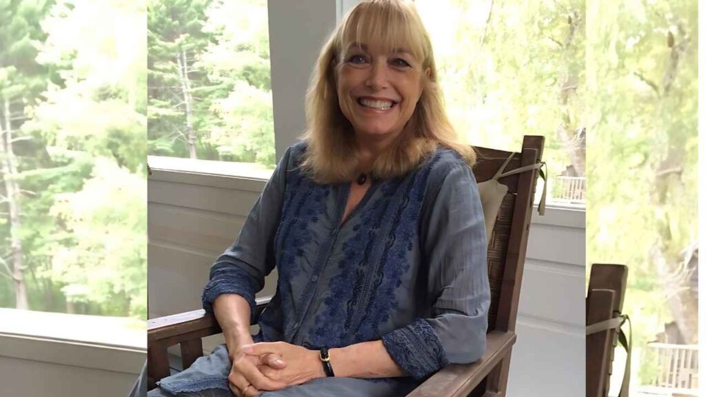 Karen Allen sits on a wooden chair beside a sunlit window, wearing a blue embroidered blouse.