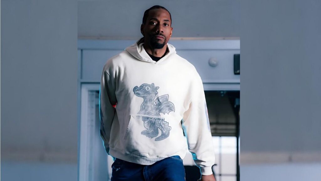 Kawhi Leonard in a white hoodie with a blue dragon graphic, standing indoors in cool, muted lighting