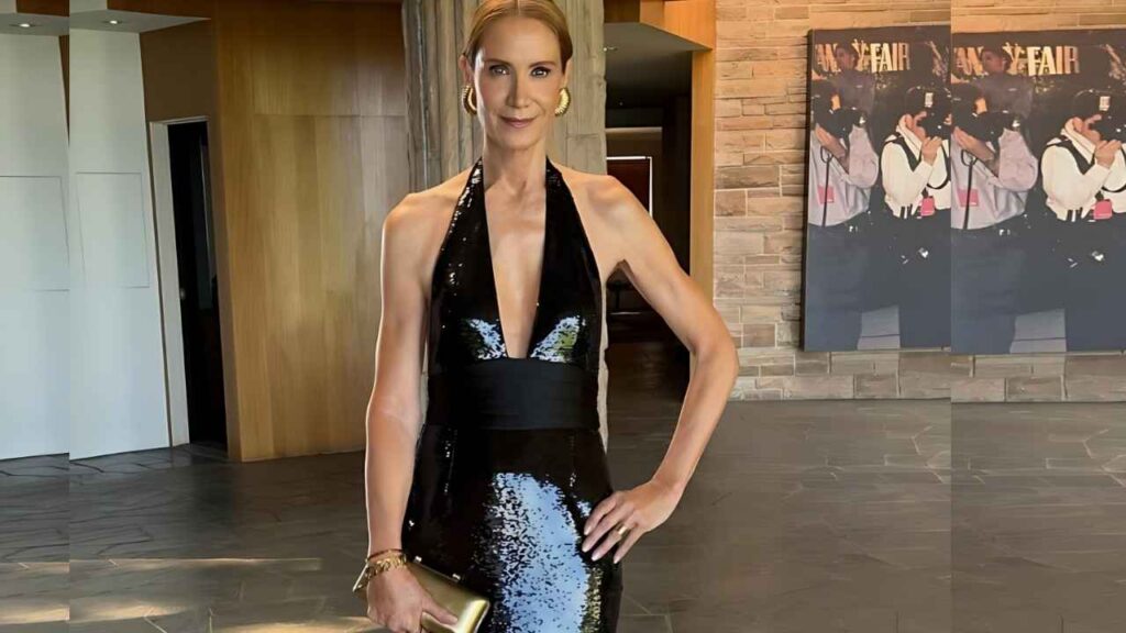 Kelly Lynch in a black sequined halter gown with a deep plunge poses in a sleek lobby, holding a gold clutch and hoops.