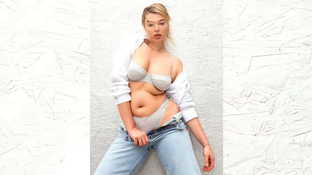 Khrystyana Kazakova in gray and a white shirt draped off her shoulders, jeans unbuttoned, posing against a white wall.