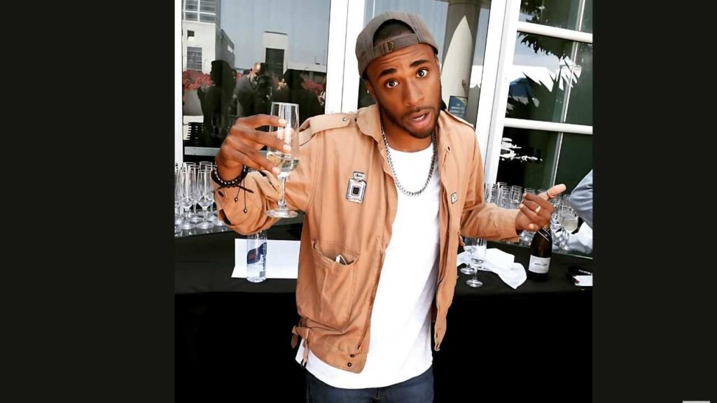 Khylin Rhambo in tan jacket and white tee, backward cap, silver chain, raises a champagne flute beside a table of glasses and a bottle.