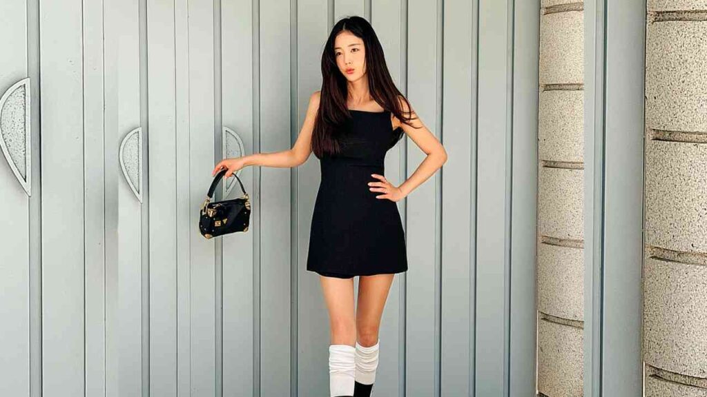 Ki Eun-se in a black mini dress poses against grey panels, holding a black handbag; white leg warmers complete her look.