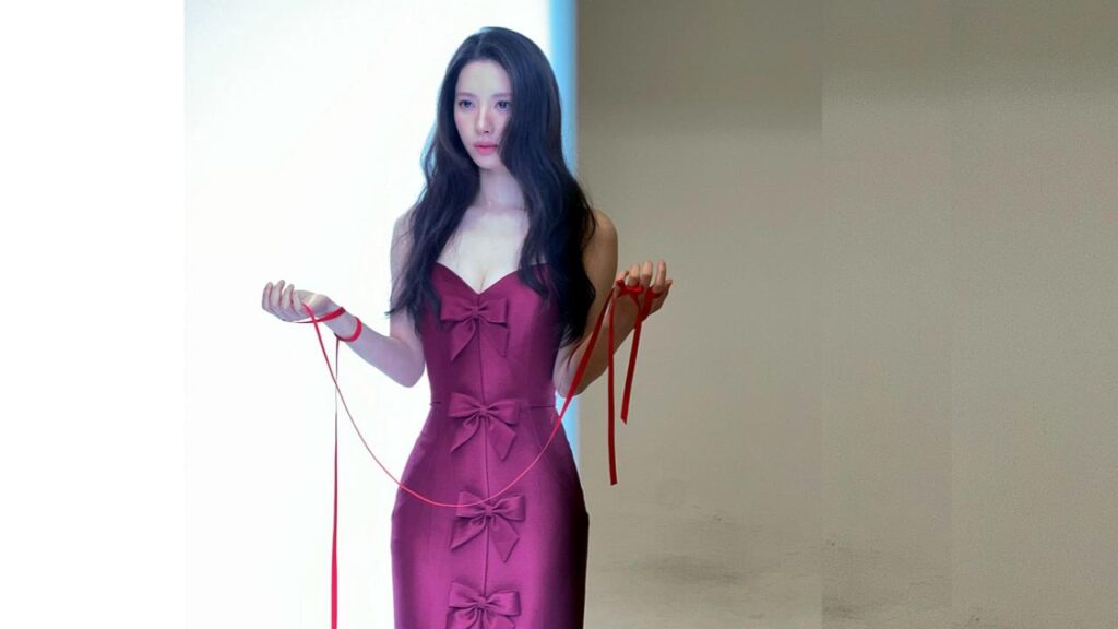 Kim Ji-yeon in a magenta strapless dress with bows along the front, holding red ribbons in each hand against a pale wall.