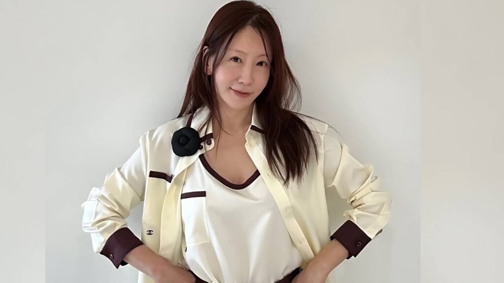 Kim Jung-eun with brown hair stands against wall in a cream jacket with dark cuffs, smiling, hands on hips, mic clipped to chest.
