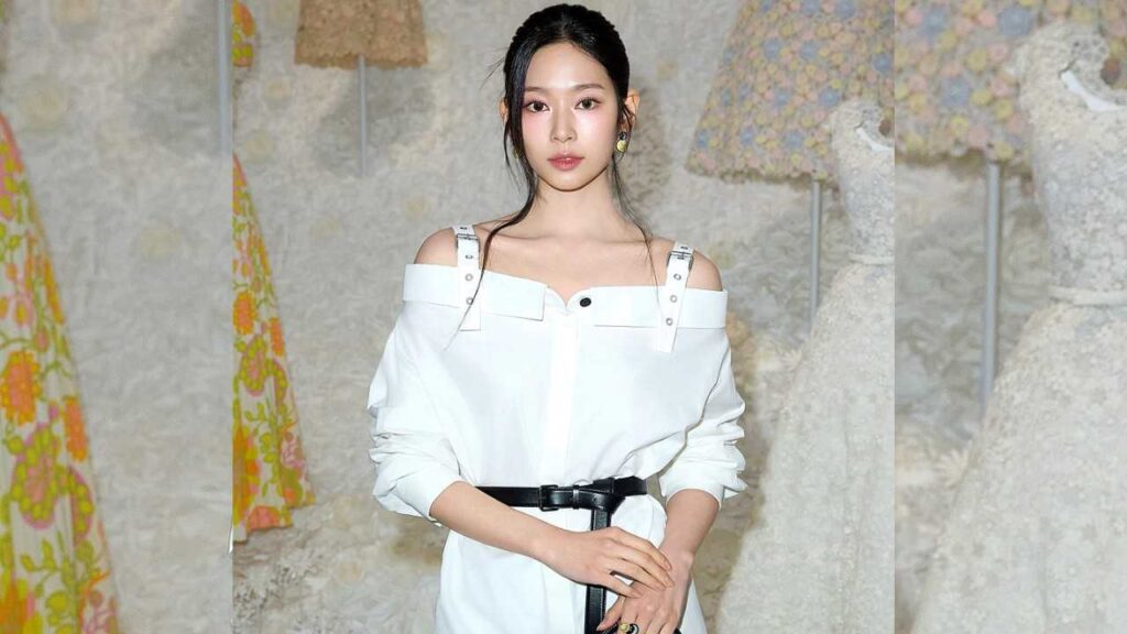 Kim Min-joo in a white off-shoulder dress with straps and a black belt, hands clasped in front, against a stone wall.