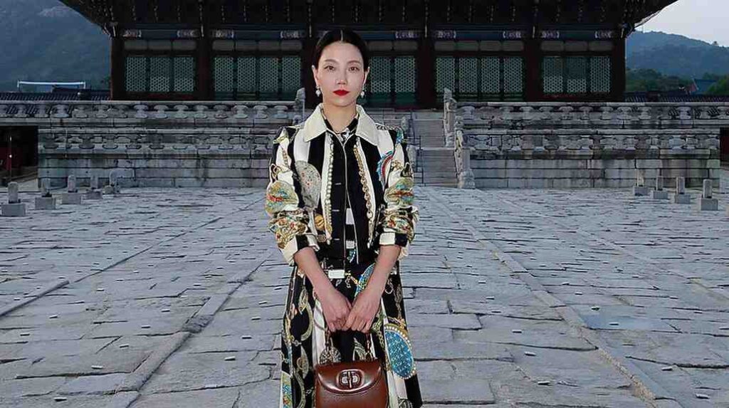 Kim Shin-rok in a vast stone courtyard before a Korean pavilion, wearing a patterned black coat and holding a brown handbag.