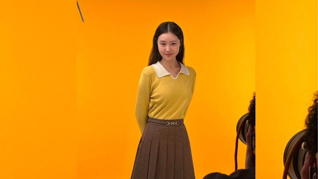 Kim Yewon (김예원) in a yellow knit sweater with white collar and a brown belted, pleated skirt, smiling.