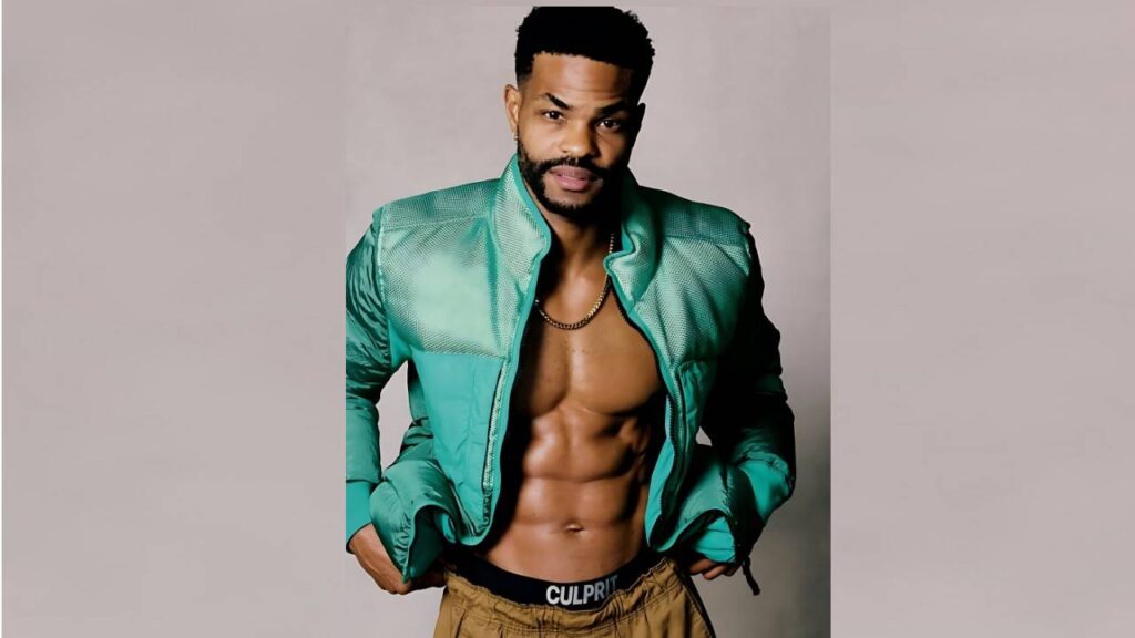 King Bach in a turquoise-green puffer jacket, open to reveal abs, wearing a gold chain, against a neutral backdrop.