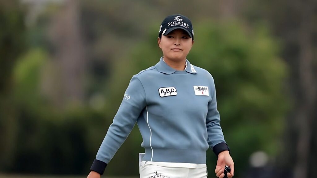 Ko Jin Young in a gray-blue pullover and white pants on a golf course, wearing a black SOLAIRE cap and patches on the chest.