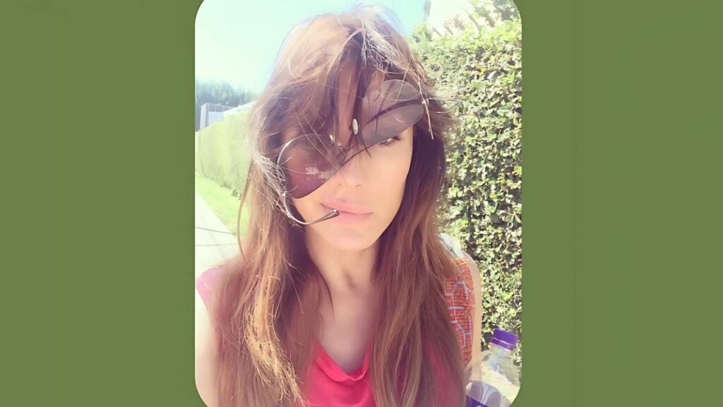 Krista Allen with long brown hair and oversized brown sunglasses outdoors, pink top, near a hedge on a sunny day.