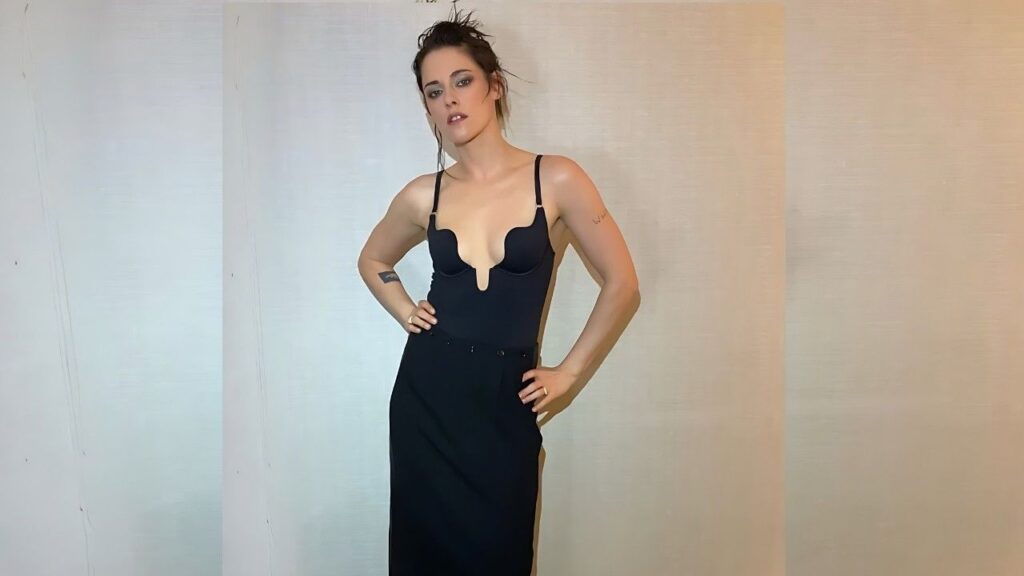 Kristen Stewart in a black plunge-neck dress stands with hands on hips, against pale panels, tousled updo and smoky makeup.