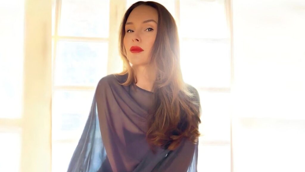 Kristina Anapau with long wavy hair and bold red lips, wearing a sheer dark top, softly lit by bright window light.