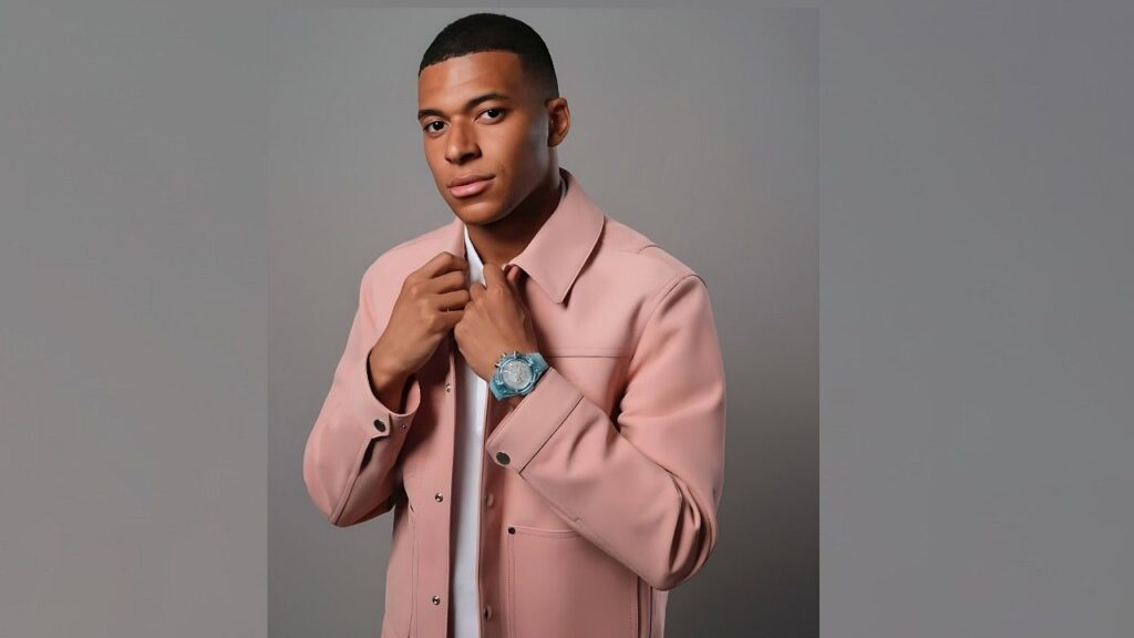 Kylian Mbappé in a pale pink jacket adjusts his lapels, white shirt underneath, turquoise wristwatch on a gray studio backdrop.