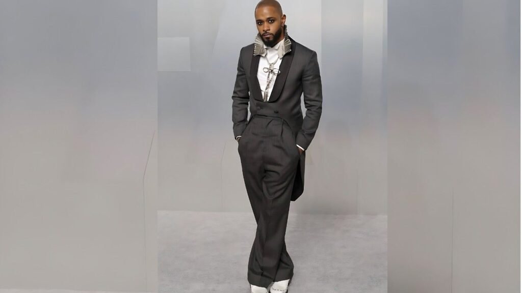 LaKeith Stanfield in a dark tailored suit with a gray ruffled collar, white shirt, and chain, hands in pockets against a gray backdrop.