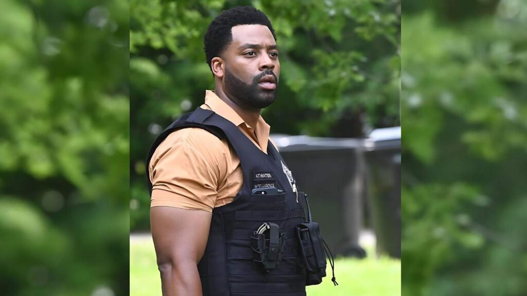 LaRoyce Hawkins wearing a black tactical vest over a tan shirt stands outdoors among lush green trees, glancing to the left.