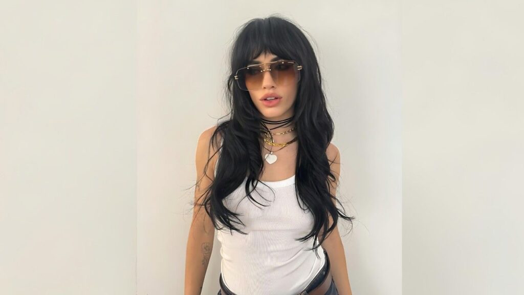 Lali Esposit with long black hair, tinted brown oversized sunglasses, white tank top, and necklaces posing against backdrop.