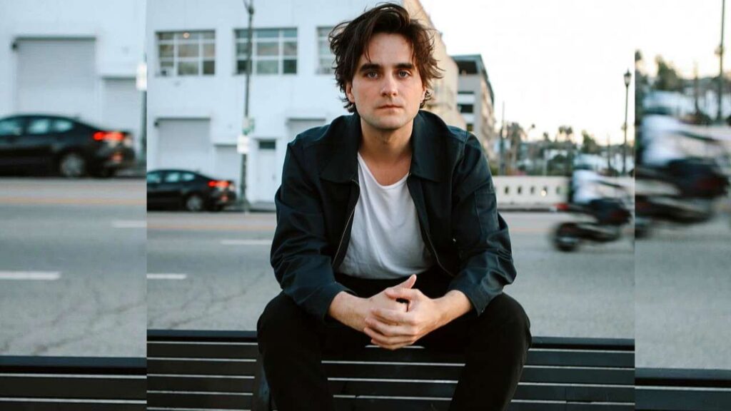 Landon Liboiron sits on a black bench by the street, hands folded, wearing a white tee and dark jacket, blurred cars behind.