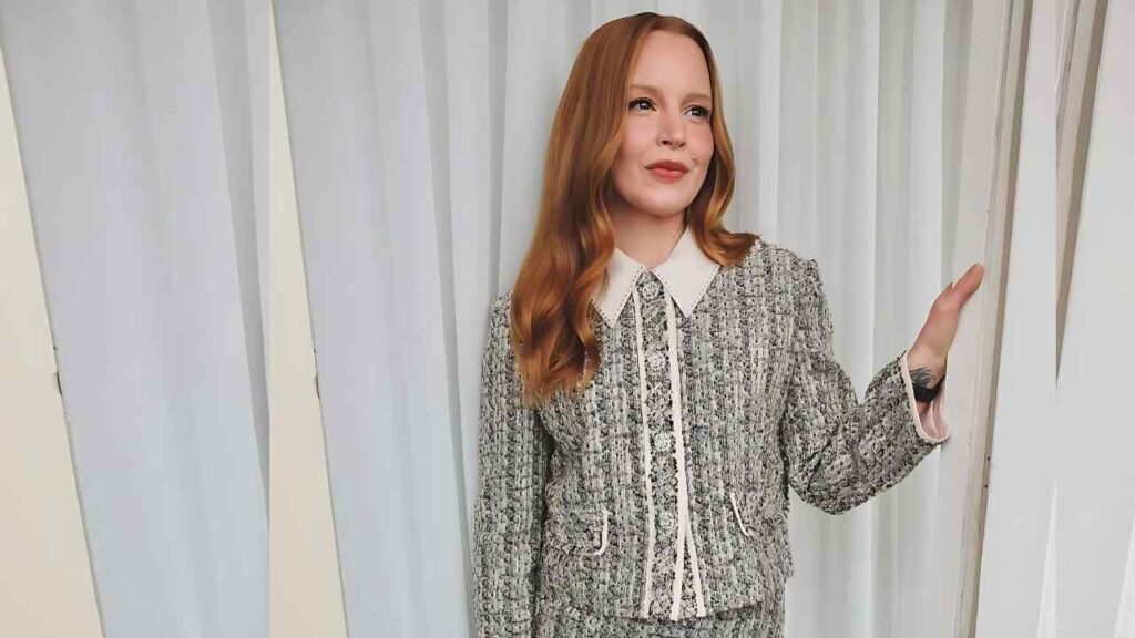 Lauren Ambrose in a textured gray tweed suit with a white collar and piping, posing beside white curtains with a soft smile.