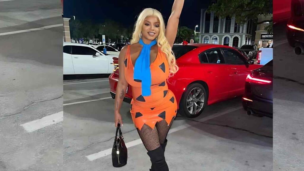 Lavish Styles in orange mini dress with black triangles and blue scarf poses in a night parking lot beside a red sports car.