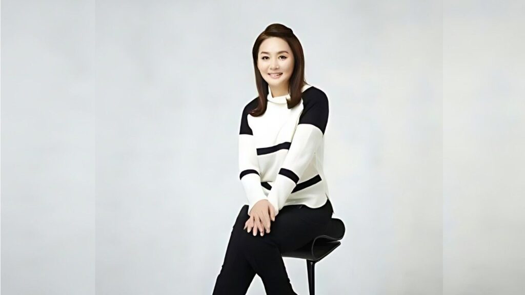 Lee Eung-kyung sits on a sleek black chair against a pale studio backdrop, wearing a white and black color-block sweater, hands folded in her lap, smiling.