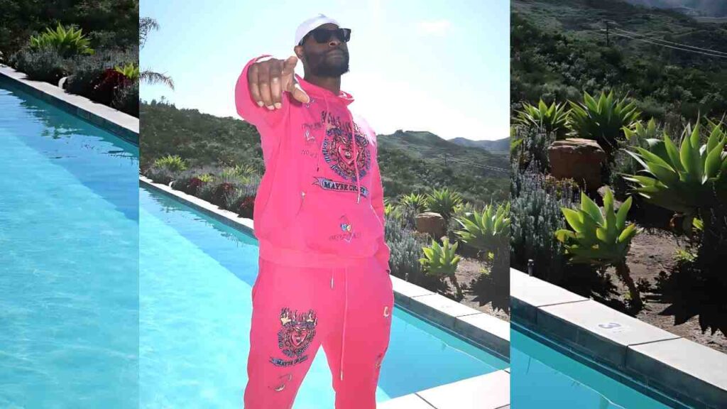 Leveon Bell in bright pink tracksuit and white cap points at the camera beside a turquoise pool, with hills in the background.