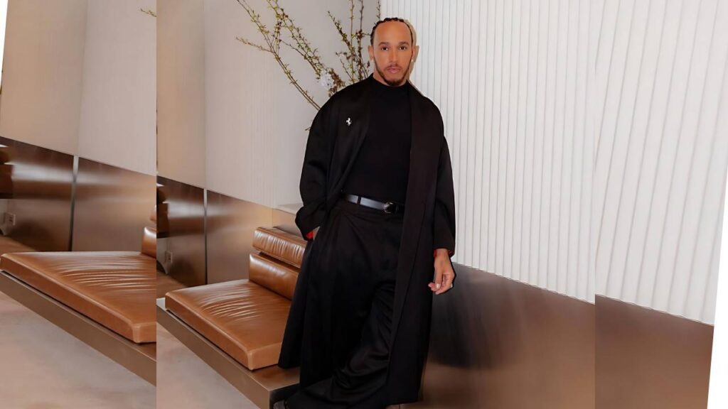 Lewis Hamilton in a long black coat and all-black outfit stands with a hand in his pocket in a modern lounge with tan leather benches.