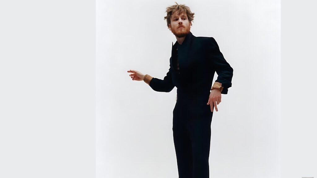 Louis Hofmann with a beard in a dark tailored suit strikes a dynamic pose in a white studio, right hand extended forward.