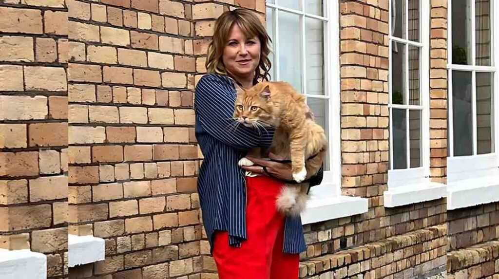 Lucy Lawless in a blue pinstriped blouse holds an orange tabby cat with white paws against a brick wall and white-framed windows.