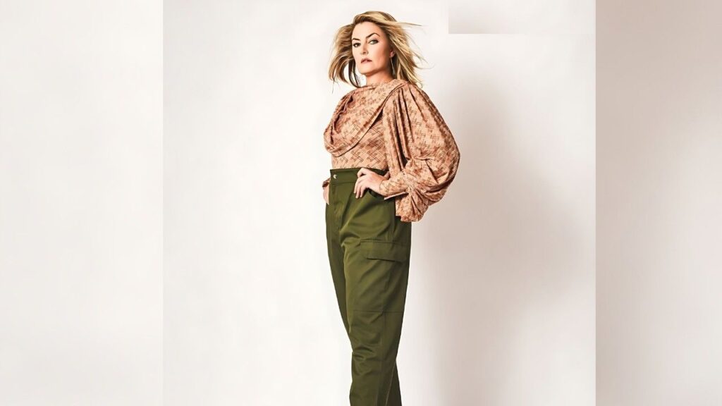 Madchen Amick stands in a neutral studio, wearing a peach patterned blouse with puff sleeves and olive cargo pants, hands on hips.