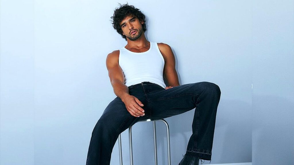 Marlon Teixeira with tousled hair sits sideways on a metal stool, wearing a white tank top and dark jeans, against a blue wall.